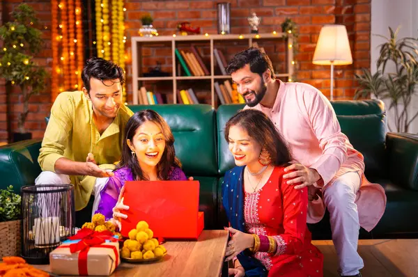 Geleneksel şenlik kıyafetleri içindeki Hintli Hindu çift, bir arkadaştan Diwali hediyesi alarak sürpriz bir şekilde arkadaşlarının hediyelerini açıyor ve güzel dekore edilmiş evlerinde neşeyle sarılıyorlar.