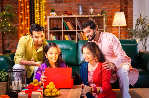 Geleneksel şenlik kıyafetleri içindeki Hintli Hindu çift, bir arkadaştan Diwali hediyesi alarak sürpriz bir şekilde arkadaşlarının hediyelerini açıyor ve güzel dekore edilmiş evlerinde neşeyle sarılıyorlar.