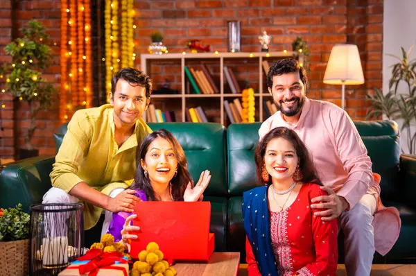 Geleneksel şenlik kıyafetleri içindeki Hintli Hindu çift, bir arkadaştan Diwali hediyesi alarak sürpriz bir şekilde arkadaşlarının hediyelerini açıyor ve güzel dekore edilmiş evlerinde neşeyle sarılıyorlar.