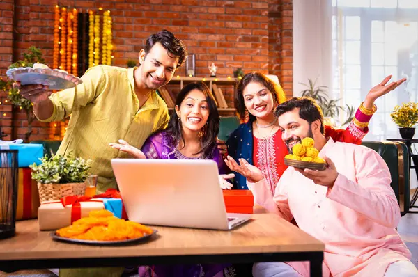 Arkadaşlar Diwali Festivali için video, Hindu Asyalı genç çift Hindu Deepavali 'yi kutlama kıyafetleri içinde dekore edilmiş evde laddu barfi tutarken dizüstü bilgisayardaki video görüşmelerine katılıyor.