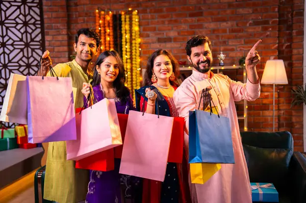 Gösterişli Diwali alışveriş torbaları ve tebessümlerle selamlaşan Hintli Asyalı genç evli çiftler dekore edilmiş evlerin iç kısımlarında geleneksel şenlik kıyafetleri içinde ve kameraya neşe dolu alışveriş torbaları gösteriyorlar.