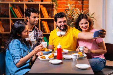 Kafede samosa ve chai içerken selfie çeken Hintli kolej öğrencileri, arkadaşlarıyla birlikte eğlenceli grup fotoğrafları çekerken aynı zamanda kapalı bir restoranda arkadaşlarıyla birlikte akıllı telefon kullanıyorlar.