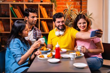 Kafede samosa ve chai içerken selfie çeken Hintli kolej öğrencileri, arkadaşlarıyla birlikte eğlenceli grup fotoğrafları çekerken aynı zamanda kapalı bir restoranda arkadaşlarıyla birlikte akıllı telefon kullanıyorlar.
