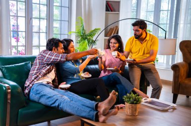 Arkadaşlar evde çayın tadını çıkarıyorlar, Hintli Asyalı genç yakın arkadaşlar sıcak çay ya da kahve içerken rahat modern evdeki kanepede dinleniyorlar sıcak bir misafirperverlik göstermek için kameraya gülümsüyorlar ve fincanları tutuyorlar.