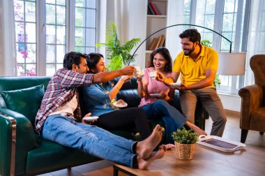 Arkadaşlar evde çayın tadını çıkarıyorlar, Hintli Asyalı genç yakın arkadaşlar sıcak çay ya da kahve içerken rahat modern evdeki kanepede dinleniyorlar sıcak bir misafirperverlik göstermek için kameraya gülümsüyorlar ve fincanları tutuyorlar.