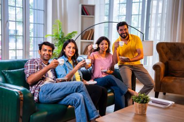 Arkadaşlar evde çayın tadını çıkarıyorlar, Hintli Asyalı genç yakın arkadaşlar sıcak çay ya da kahve içerken rahat modern evdeki kanepede dinleniyorlar sıcak bir misafirperverlik göstermek için kameraya gülümsüyorlar ve fincanları tutuyorlar.