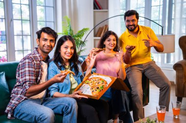 Hintli arkadaşlar pizza yiyorlar ve derin sohbetlere dalıyorlar, Asyalı genç yakın arkadaşlar modern evdeki rahat koltukta oturuyorlar, yemeğin tadını çıkarıyorlar, kaynaşıyorlar, gülüyorlar, düşüncelerini paylaşıyorlar ve evde dinleniyorlar.