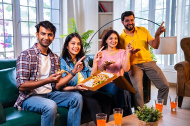 Hintli arkadaşlar pizza yiyorlar ve derin sohbetlere dalıyorlar, Asyalı genç yakın arkadaşlar modern evdeki rahat koltukta oturuyorlar, yemeğin tadını çıkarıyorlar, kaynaşıyorlar, gülüyorlar, düşüncelerini paylaşıyorlar ve evde dinleniyorlar.