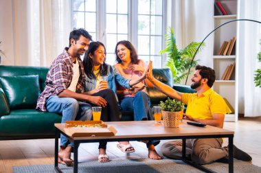 Hintli arkadaşlar kameraya gülümserken rahat bir koltukta oturup pizza yiyorlar, birlikte gündelik zaman geçiriyorlar, kaynaşıyorlar, gülüyorlar ve modern evde dinleniyorlar.