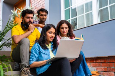 Üniversite kampüsündeki başarıyı kutlayan Hintli üniversite öğrencileri grup ödevlerini kitaplarla ve notlarla tamamlarken dışarıda birlikte oturuyorlar ve neşeli bir şekilde başparmaklarını gösteriyorlar.