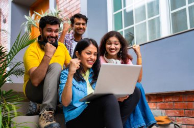 Üniversite kampüsündeki başarıyı kutlayan Hintli üniversite öğrencileri grup ödevlerini kitaplarla ve notlarla tamamlarken dışarıda birlikte oturuyorlar ve neşeli bir şekilde başparmaklarını gösteriyorlar.