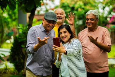 Son sınıf arkadaşlarının videosu akıllı telefondan arıyor, neşeli Hintli Asyalı yaşlı grup açık havada yemyeşil bahçede duruyorlar, el sallıyorlar, sevdikleriyle mobil video iletişimi sırasında gülümsüyorlar.