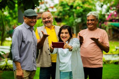 Yaşlılar akıllı telefondan fotoğraflara bakıyor ve gülümsüyor, Hintli Asyalı yaşlılar bahçe grubunda duruyorlar, gezici fotoğraf galerilerini izliyorlar, dışarıda yeşil yapraklarla çevrili iken sevinçlerini ifade ediyorlar.
