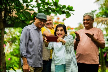 Yaşlılar akıllı telefondan fotoğraflara bakıyor ve gülümsüyor, Hintli Asyalı yaşlılar bahçe grubunda duruyorlar, gezici fotoğraf galerilerini izliyorlar, dışarıda yeşil yapraklarla çevrili iken sevinçlerini ifade ediyorlar.