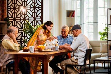 Son sınıflar birlikte yemek yiyor, Hintli Asyalı yaşlı arkadaşlar evde bir araya geliyor, yemek yiyor, atıştırmalık, soğuk içecek, çay ya da kahve servisi yapıyor, modern yemek masasında oturmuş, eğleniyorlar.