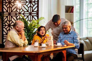 Hint kıdemli yetişkinler, Hint evinde yemek masasında oturmuş, gülüyor, eğlenceli anları kutluyor, kaynaşıyor, arkadaşça bir araya gelip eğleniyorlar.