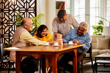 Hint kıdemli yetişkinler, Hint evinde yemek masasında oturmuş, gülüyor, eğlenceli anları kutluyor, kaynaşıyor, arkadaşça bir araya gelip eğleniyorlar.
