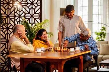 Hindistan 'daki son sınıf yetişkinler rahat bir evde yemek masasında oturmuş satranç oynarken gülümsüyor ve Shatranj maçının keyfini çıkarırken Asyalı eski dostlar kapalı alanda yeniden bir araya gelişlerini kutluyorlar.