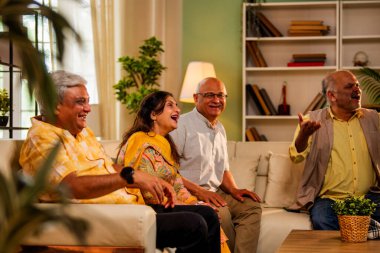 Hintli son sınıf öğrencisi rahat bir koltukta oturup televizyon izlerken gülerken Asyalı yaşlı erkekler ve kadınlar ellerinde uzaktan kumandayla televizyon izlerken televizyonda komik film ya da komedi şovu izliyorlar.