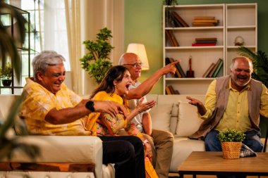 Hintli son sınıf öğrencisi rahat bir koltukta oturup televizyon izlerken gülerken Asyalı yaşlı erkekler ve kadınlar ellerinde uzaktan kumandayla televizyon izlerken televizyonda komik film ya da komedi şovu izliyorlar.