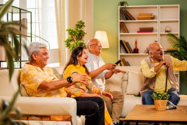 Hintli son sınıf öğrencisi rahat bir koltukta oturup televizyon izlerken gülerken Asyalı yaşlı erkekler ve kadınlar ellerinde uzaktan kumandayla televizyon izlerken televizyonda komik film ya da komedi şovu izliyorlar.