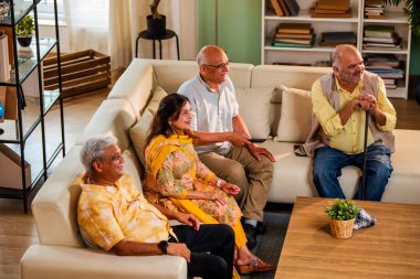 Hintli son sınıf öğrencisi rahat bir koltukta oturup televizyon izlerken gülerken Asyalı yaşlı erkekler ve kadınlar ellerinde uzaktan kumandayla televizyon izlerken televizyonda komik film ya da komedi şovu izliyorlar.