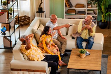 Hintli son sınıf öğrencisi rahat bir koltukta oturup televizyon izlerken gülerken Asyalı yaşlı erkekler ve kadınlar ellerinde uzaktan kumandayla televizyon izlerken televizyonda komik film ya da komedi şovu izliyorlar.