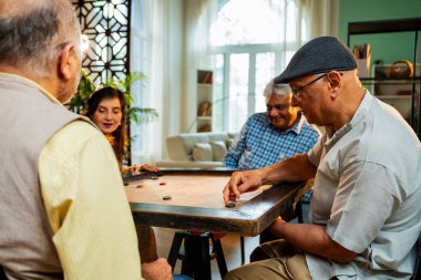Hintli son sınıflar birlikte karrom masası oyunu oynuyorlar, rahat bir evde yemek masasında oturuyorlar, arkadaşça bir maç izliyorlar, gülüyorlar, emekli Hintli erkek ve kadınlar eğlenceli buluşma aktivitelerini kutluyorlar.
