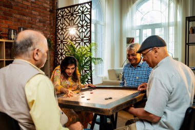 Hintli son sınıflar birlikte karrom masası oyunu oynuyorlar, rahat bir evde yemek masasında oturuyorlar, arkadaşça bir maç izliyorlar, gülüyorlar, emekli Hintli erkek ve kadınlar eğlenceli buluşma aktivitelerini kutluyorlar.