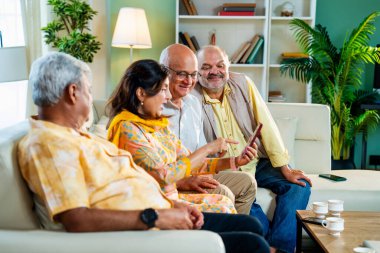 Hint Asya modern evindeki rahat koltukta otururken akıllı telefonları kontrol eden kıdemli arkadaşlar, rahat, neşeli ve mutlu bir şekilde cep telefonuyla buluşurken fotoğraflara bakıp gülüyorlar.