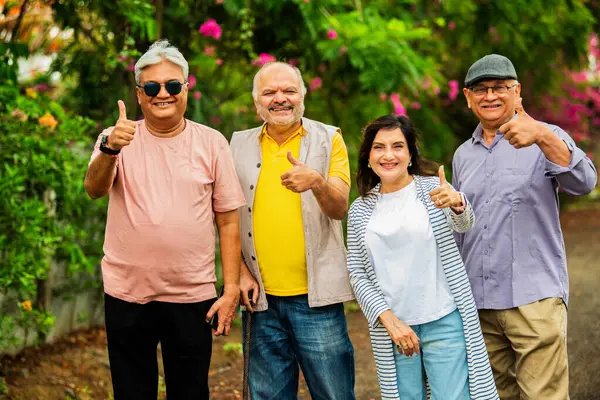 Yaşlı yetişkinler neşeli gülümsemelerle başparmak gösteriyorlar, Hintli Asyalı yaşlı arkadaşlar çiçeklerle dolu yeşil bahçeye karşı bir grupta duruyorlar, kameraya bakarken pozitif jestler yapıyorlar.