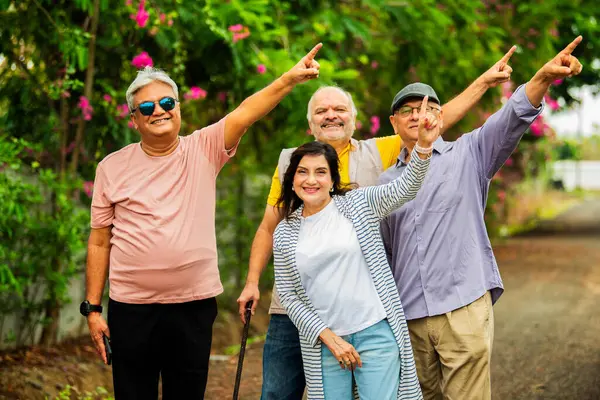 Hintli kıdemli yetişkinler dışarıyı gösteriyor ve gülümsüyor, dört mutlu Asyalı yaşlı arkadaş çiçeklerle dolu yemyeşil bir arka plana karşı grup halinde duruyorlar, gülümsüyorlar ve bir şey sunuyorlar ya da sunuyorlar.