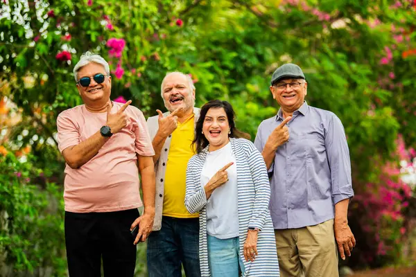Hintli kıdemli yetişkinler dışarıyı gösteriyor ve gülümsüyor, dört mutlu Asyalı yaşlı arkadaş çiçeklerle dolu yemyeşil bir arka plana karşı grup halinde duruyorlar, gülümsüyorlar ve bir şey sunuyorlar ya da sunuyorlar.
