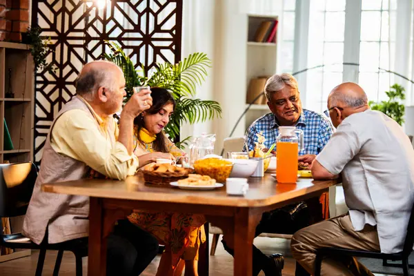 Son sınıflar yemek yerken sohbet ediyorlar, Hintli Asyalı yaşIı arkadaşlar yemek masasının etrafında neşeli sohbetler ve kahkahalar atarak evde atıştırmalık, çay, kahve ve yemek paylaşıyorlar.