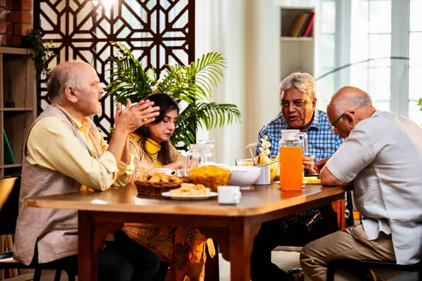 Son sınıflar yemek yerken sohbet ediyorlar, Hintli Asyalı yaşIı arkadaşlar yemek masasının etrafında neşeli sohbetler ve kahkahalar atarak evde atıştırmalık, çay, kahve ve yemek paylaşıyorlar.