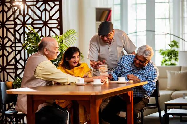 Hint kıdemli yetişkinler, Hint evinde yemek masasında oturmuş, gülüyor, eğlenceli anları kutluyor, kaynaşıyor, arkadaşça bir araya gelip eğleniyorlar.