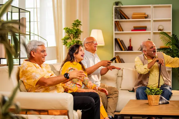 Hintli son sınıf öğrencisi rahat bir koltukta oturup televizyon izlerken gülerken Asyalı yaşlı erkekler ve kadınlar ellerinde uzaktan kumandayla televizyon izlerken televizyonda komik film ya da komedi şovu izliyorlar.