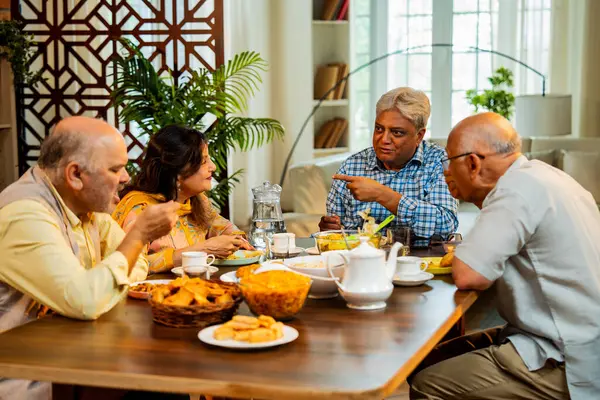 Son sınıflar yemek yerken sohbet ediyorlar, Hintli Asyalı yaşIı arkadaşlar yemek masasının etrafında neşeli sohbetler ve kahkahalar atarak evde atıştırmalık, çay, kahve ve yemek paylaşıyorlar.