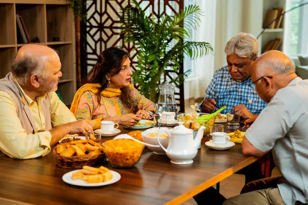 Son sınıflar yemek yerken sohbet ediyorlar, Hintli Asyalı yaşIı arkadaşlar yemek masasının etrafında neşeli sohbetler ve kahkahalar atarak evde atıştırmalık, çay, kahve ve yemek paylaşıyorlar.