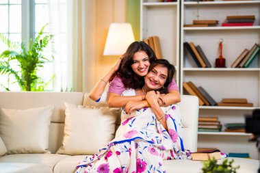 Hintli kıdemli anne kızı koltukta sarılıyor. Asyalı genç kız yaşlı anneye sürpriz yapıyor. Kitap okuyor, hem sohbet ediyor hem de modern oturma odasında mutlu bir bağ kuruyor.