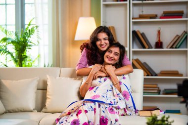 Hintli kıdemli anne kızı koltukta sarılıyor. Asyalı genç kız yaşlı anneye sürpriz yapıyor. Kitap okuyor, hem sohbet ediyor hem de modern oturma odasında mutlu bir bağ kuruyor.