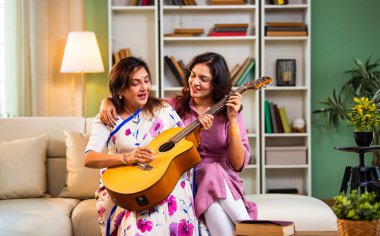 Anne kızından gitar öğreniyor, Hintli yaşlı anne kanepede oturuyor genç kızından müzik dersi alıyor modern evde neşeyle akustik gitar çalıyor.
