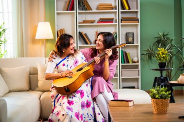 Anne kızından gitar öğreniyor, Hintli yaşlı anne kanepede oturuyor genç kızından müzik dersi alıyor modern evde neşeyle akustik gitar çalıyor.