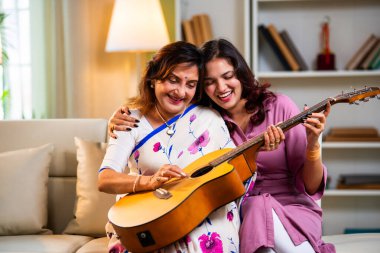 Anne kızından gitar öğreniyor, Hintli yaşlı anne kanepede oturuyor genç kızından müzik dersi alıyor modern evde neşeyle akustik gitar çalıyor.