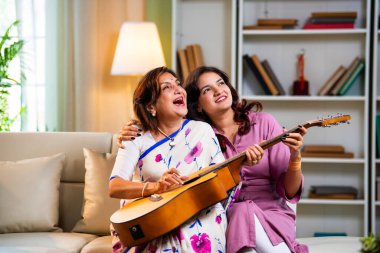 Anne kızından gitar öğreniyor, Hintli yaşlı anne kanepede oturuyor genç kızından müzik dersi alıyor modern evde neşeyle akustik gitar çalıyor.