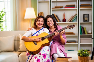 Anne kızından gitar öğreniyor, Hintli yaşlı anne kanepede oturuyor genç kızından müzik dersi alıyor modern evde neşeyle akustik gitar çalıyor.