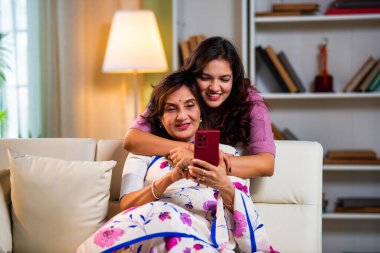 Yaşlı anne kız akıllı telefon kullanıyor, Hintli Asya 'lı kıdemli anne ve genç kız kanepenin arkasından sarılıyor, hem parşömen, hem alışveriş hem de sörf yapıyor modern evde neşeyle dokunmatik ekranda geziyor.