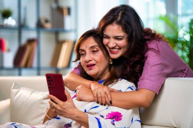 Yaşlı anne kız akıllı telefon kullanıyor, Hintli Asya 'lı kıdemli anne ve genç kız kanepenin arkasından sarılıyor, hem parşömen, hem alışveriş hem de sörf yapıyor modern evde neşeyle dokunmatik ekranda geziyor.