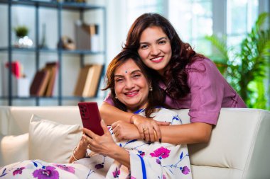 Yaşlı anne kız akıllı telefon kullanıyor, Hintli Asya 'lı kıdemli anne ve genç kız kanepenin arkasından sarılıyor, hem parşömen, hem alışveriş hem de sörf yapıyor modern evde neşeyle dokunmatik ekranda geziyor.