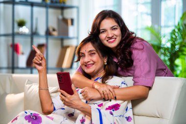 Yaşlı anne kız akıllı telefon kullanıyor, Hintli Asya 'lı kıdemli anne ve genç kız kanepenin arkasından sarılıyor, hem parşömen, hem alışveriş hem de sörf yapıyor modern evde neşeyle dokunmatik ekranda geziyor.